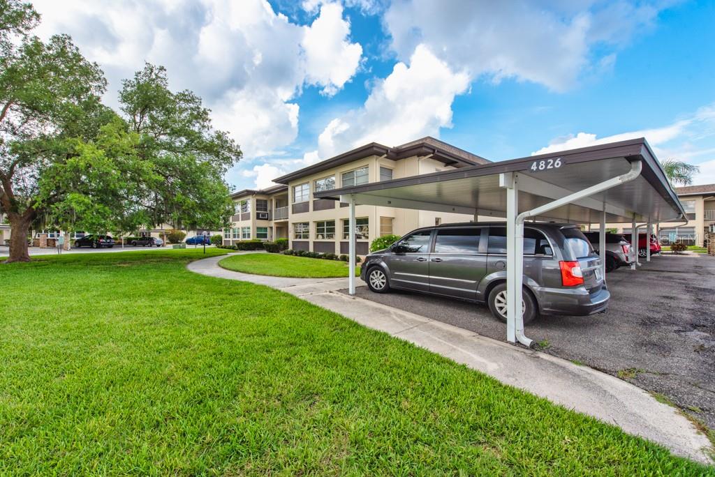 A gray minivan is parked under a covered carport next to a multi-story apartment building — 4826 MARINE PARKWAY UNIT 204, NEW PORT RICHEY, FL