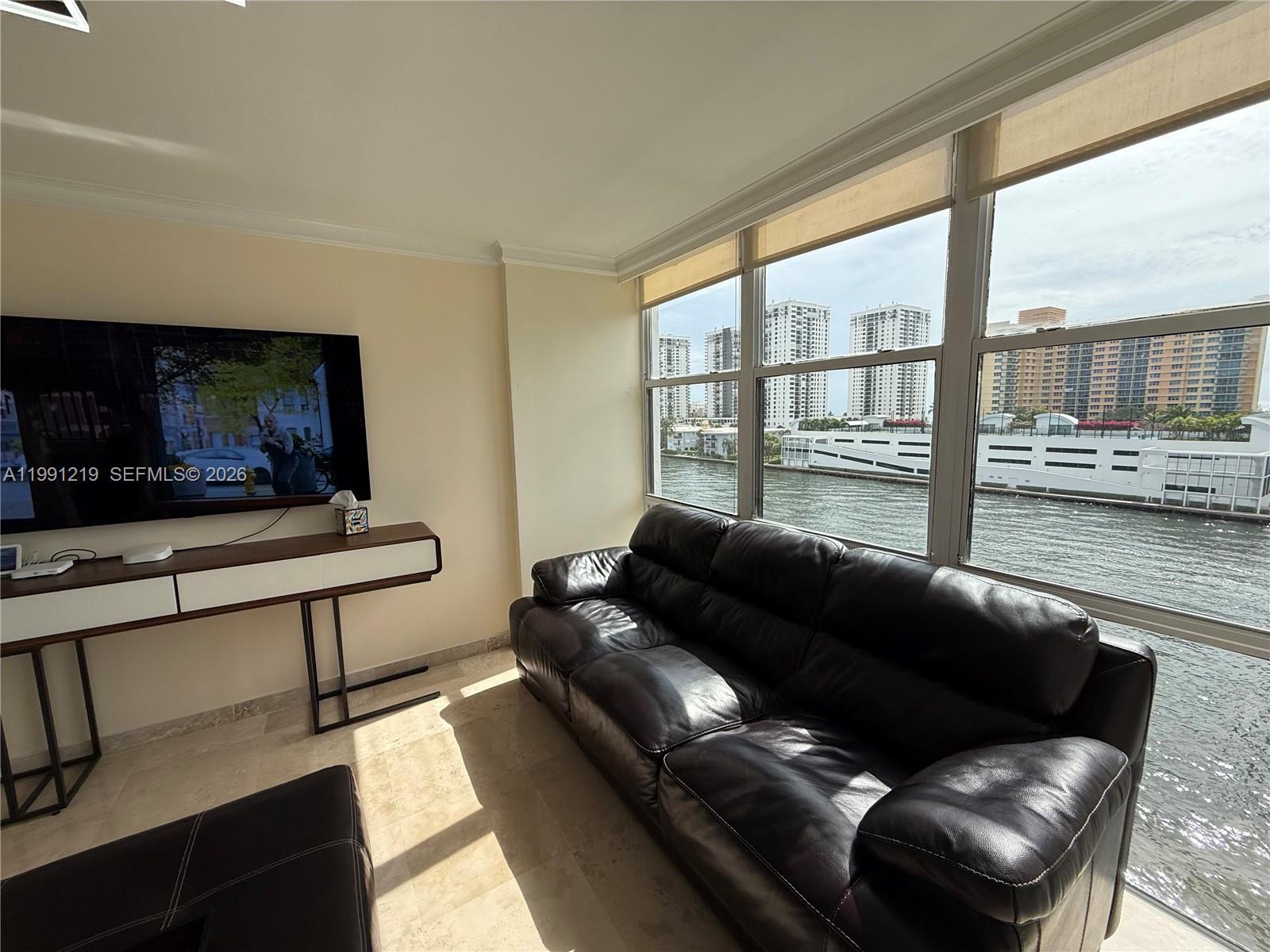 A living room features a dark leather sofa, a wall-mounted television, and floor-to-ceiling windows — 1000 Parkview Dr # 720, Hallandale Beach FL 33009, HALLANDALE BEACH, FL