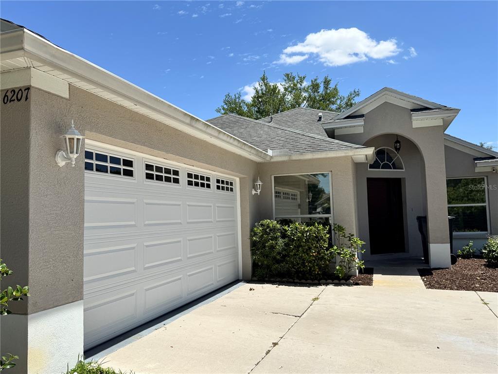 A beige single-story house exterior features a two-car garage, a front entryway, and landscaping — 6207 TUPELO TRAIL, LAKEWOOD RANCH, FL