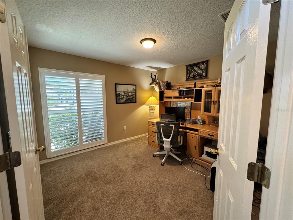 A home office features a wooden desk, an ergonomic chair, and plantation shutters on the window — 6207 TUPELO TRAIL, LAKEWOOD RANCH, FL