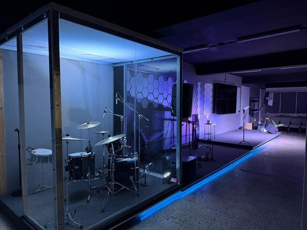 A drum kit sits inside a glass enclosure on a stage with blue accent lighting — 100 SECTOR LA CUESTA, LOS JOBOS, HUMACAO, PR