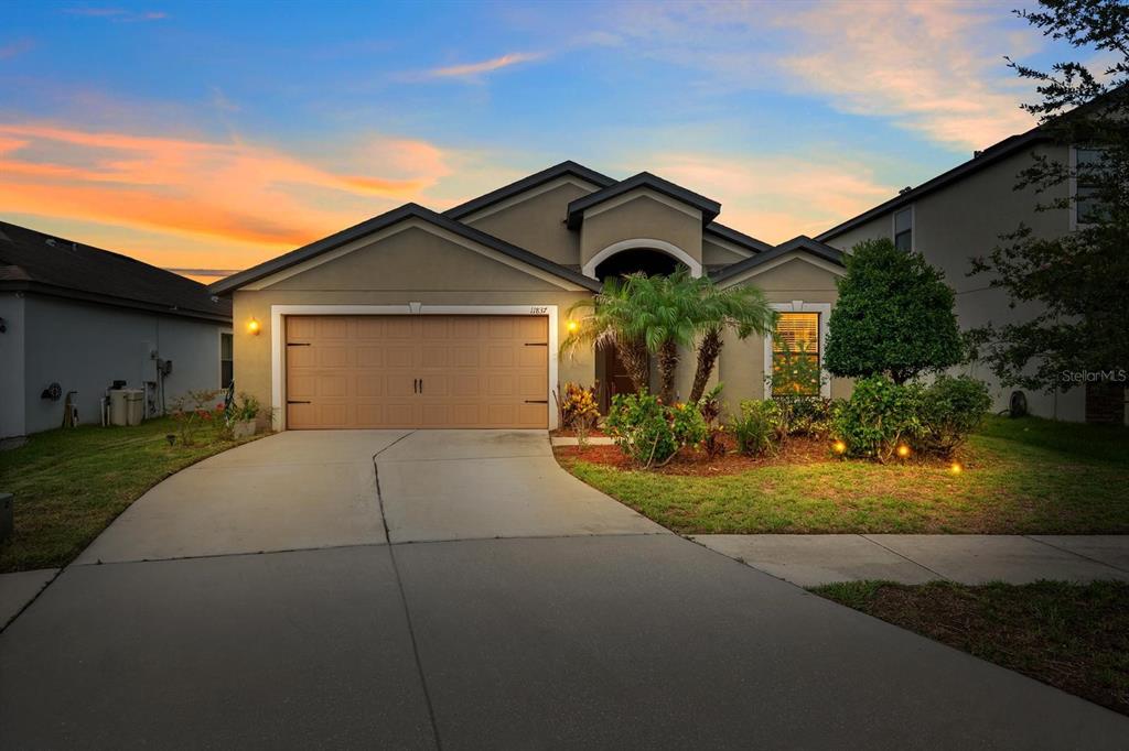 A single-story house with a tan stucco exterior, a two-car garage, and a landscaped front yard — 11837 THICKET WOOD DRIVE, RIVERVIEW, FL