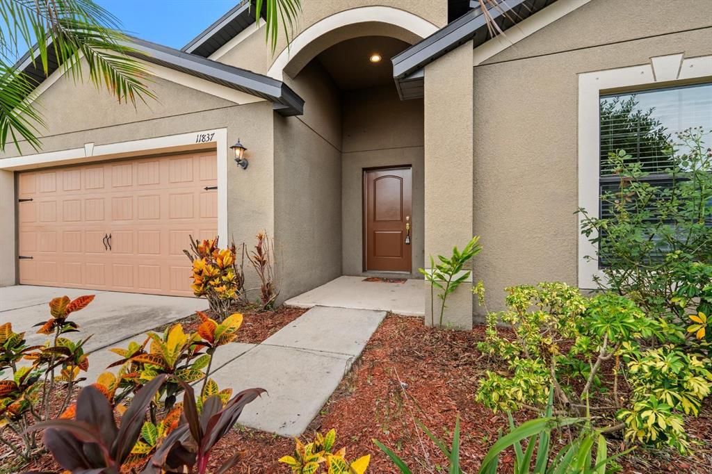A tan stucco house exterior features a brown garage door, arched entryway, and landscaped yard — 11837 THICKET WOOD DRIVE, RIVERVIEW, FL