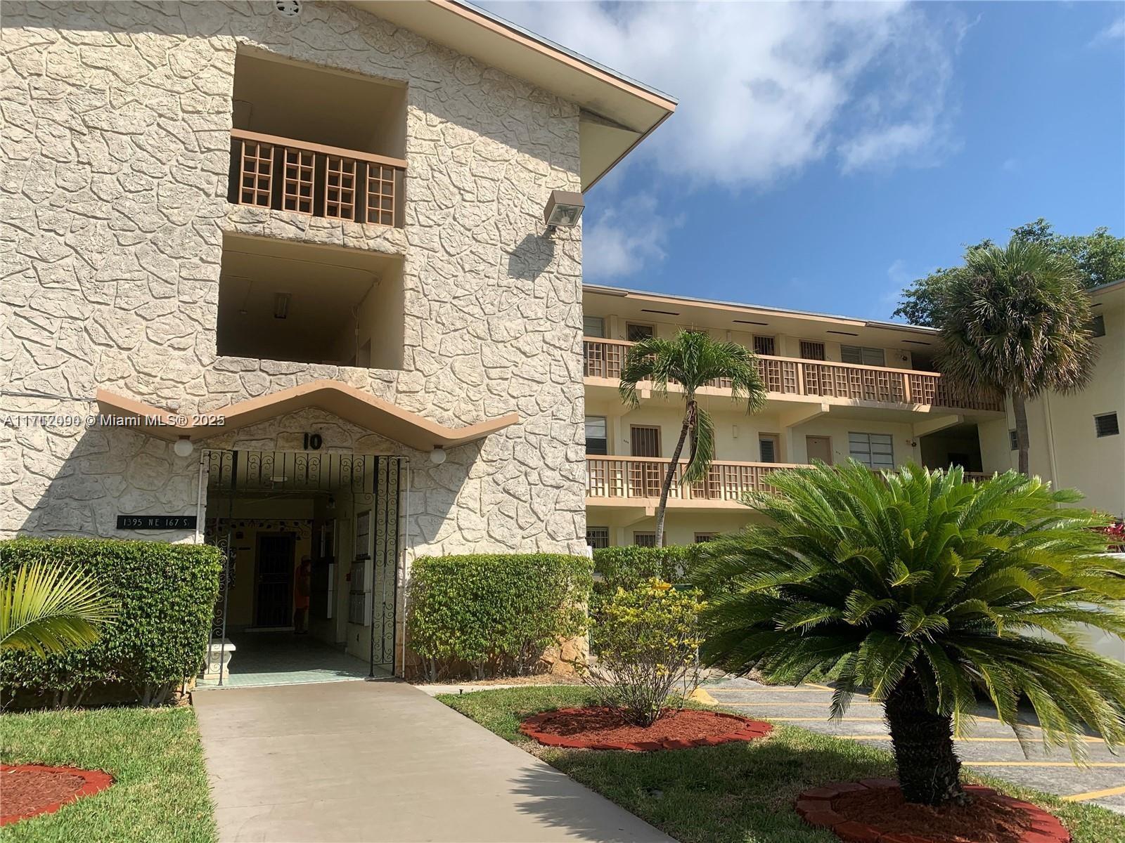 A multi-story apartment building features a stone facade, open balconies, and a palm-lined entrance — 1395 NE 167th St # 209, Miami FL 33162, MIAMI, FL