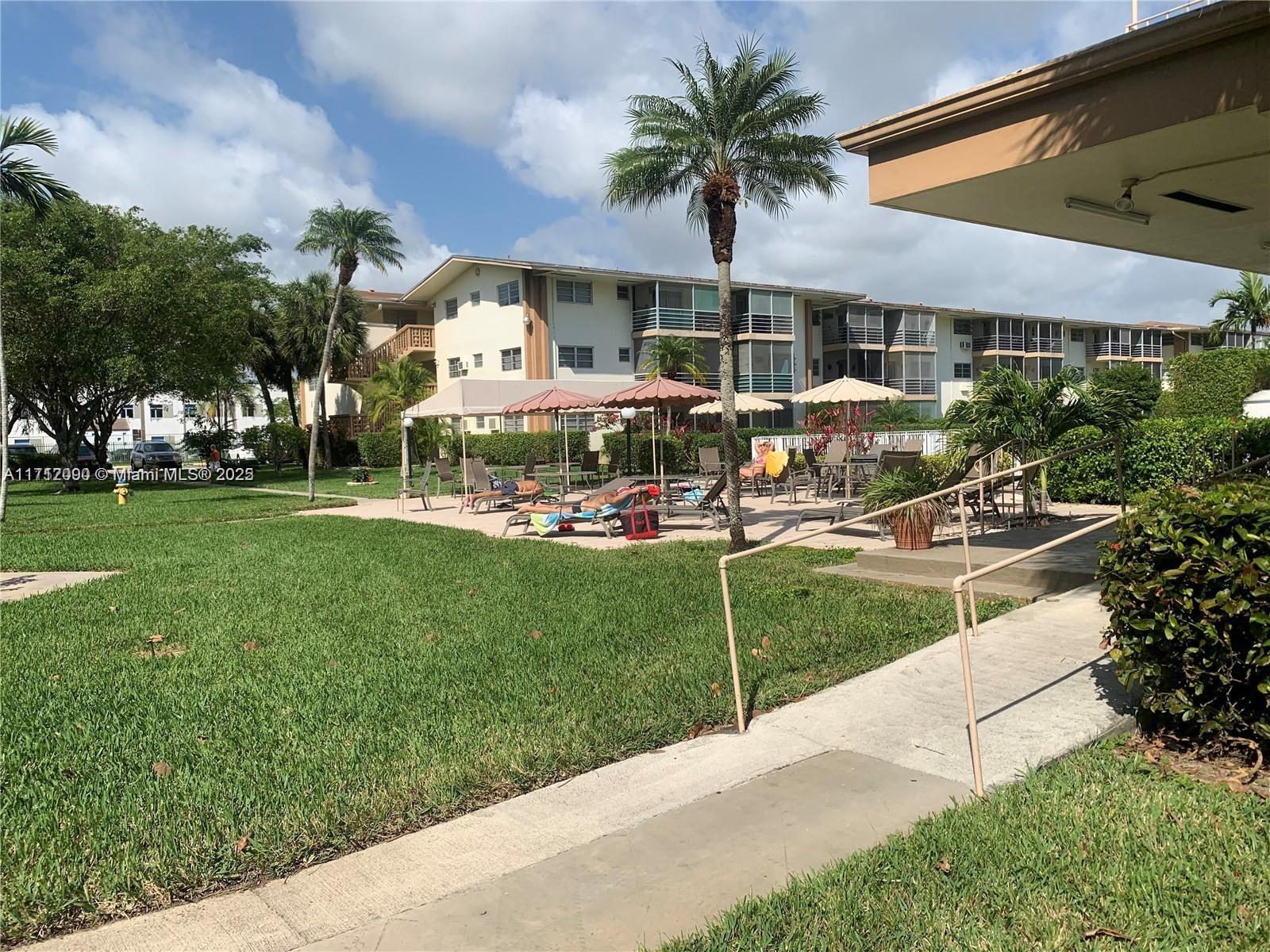 A grassy lawn leads to a patio area with lounge chairs and umbrellas near apartments — 1395 NE 167th St # 209, Miami FL 33162, MIAMI, FL