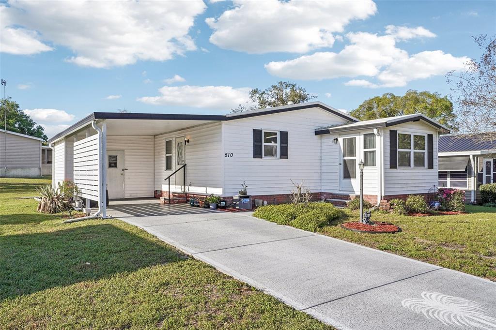 A white single-story manufactured home with a carport, concrete driveway, and a small front yard — 510 BONITA DRIVE, LADY LAKE, FL