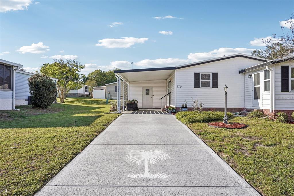 A white mobile home with a carport and a palm tree stencil on the driveway — 510 BONITA DRIVE, LADY LAKE, FL