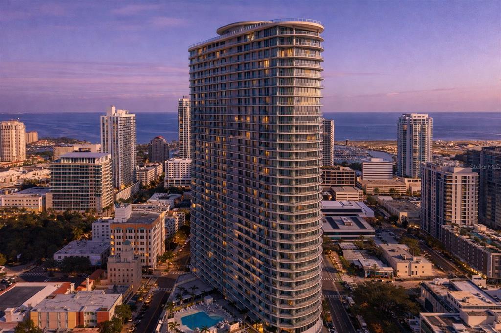 An aerial view of a tall, curved glass residential skyscraper in a coastal city — 400 CENTRAL AVENUE UNIT 1009, ST PETERSBURG, FL