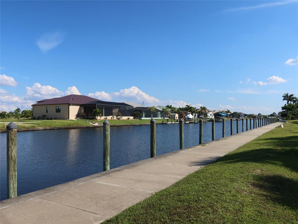 A concrete walkway runs alongside a canal lined with wooden pilings and residential homes — 8185 DIMSTEAD STREET, PORT CHARLOTTE, FL