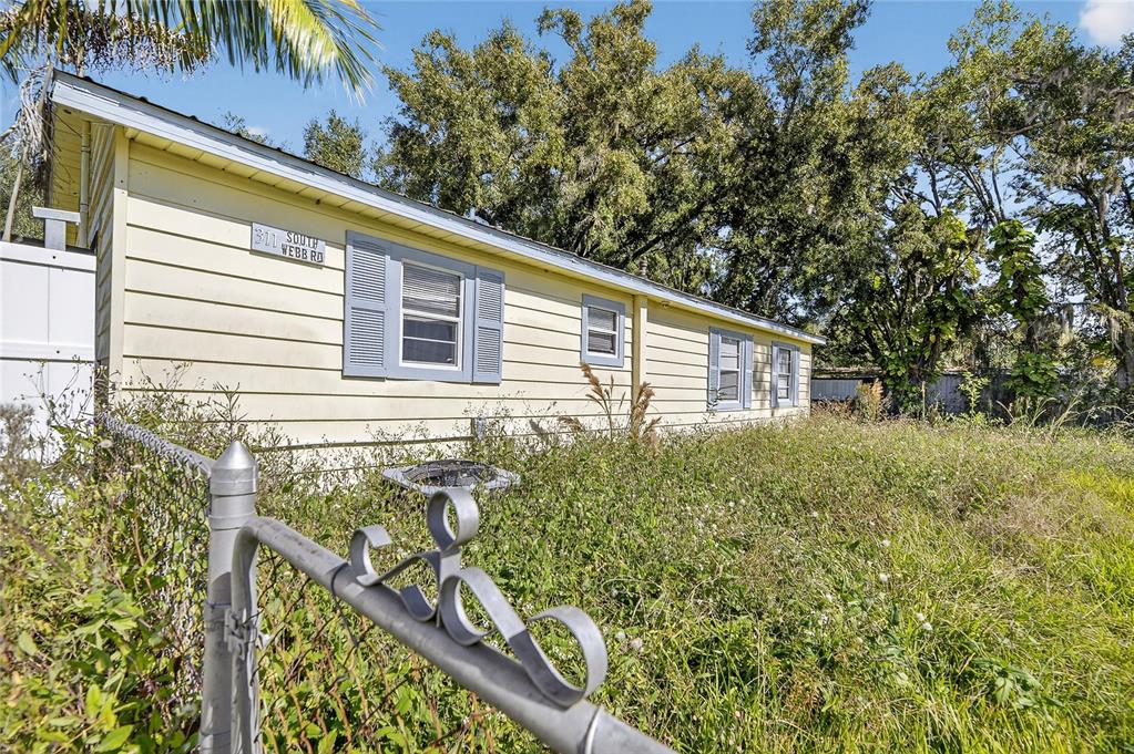 A light yellow house with blue shutters stands behind a chain-link fence and overgrown grass — 311 S WEBB ROAD, PLANT CITY, FL