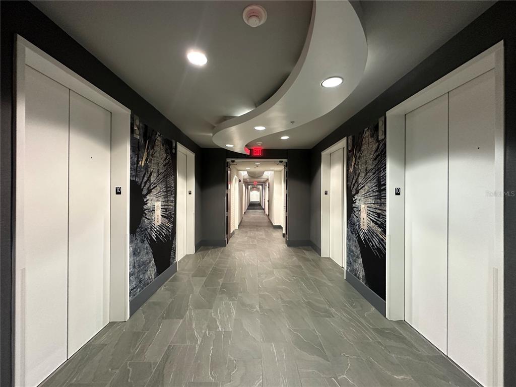 A modern apartment building hallway features white elevator doors, dark patterned walls, and tile flooring — 155 S COURT AVENUE UNIT 1911, ORLANDO, FL