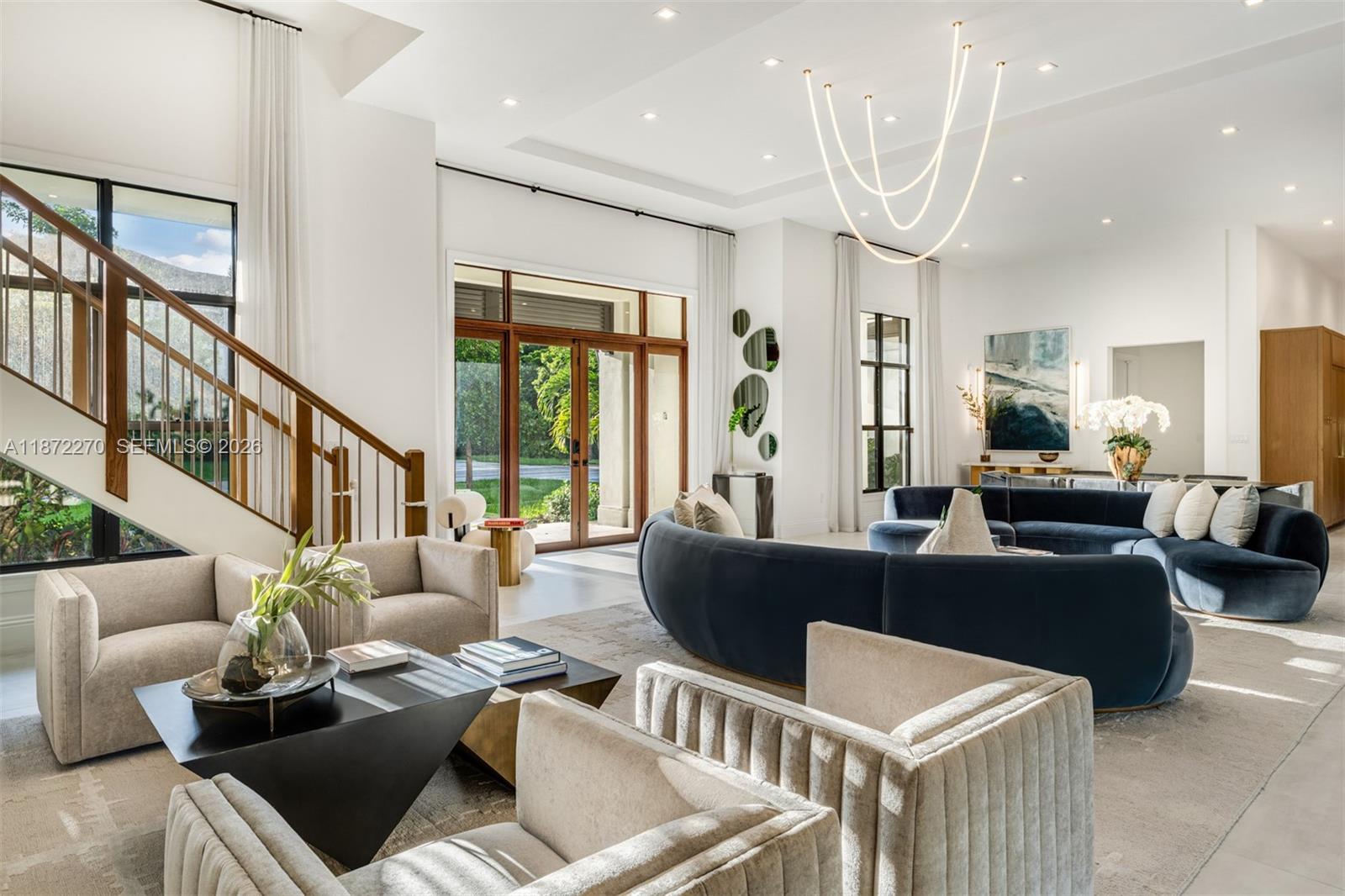 A spacious living room features a curved blue sofa, neutral armchairs, and a modern chandelier — 9445 SW 72nd Ct, Pinecrest FL 33156, PINECREST, FL