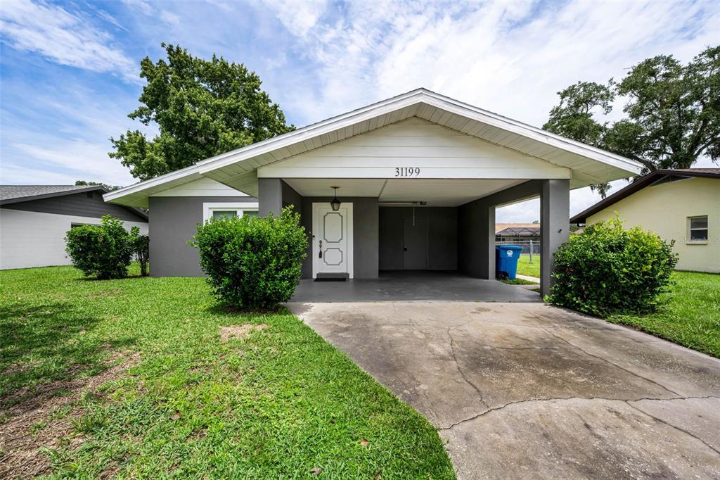 A single-story gray house features a front carport, a white front door, and green landscaping — 31199 LANCEWOOD DRIVE, BROOKSVILLE, FL