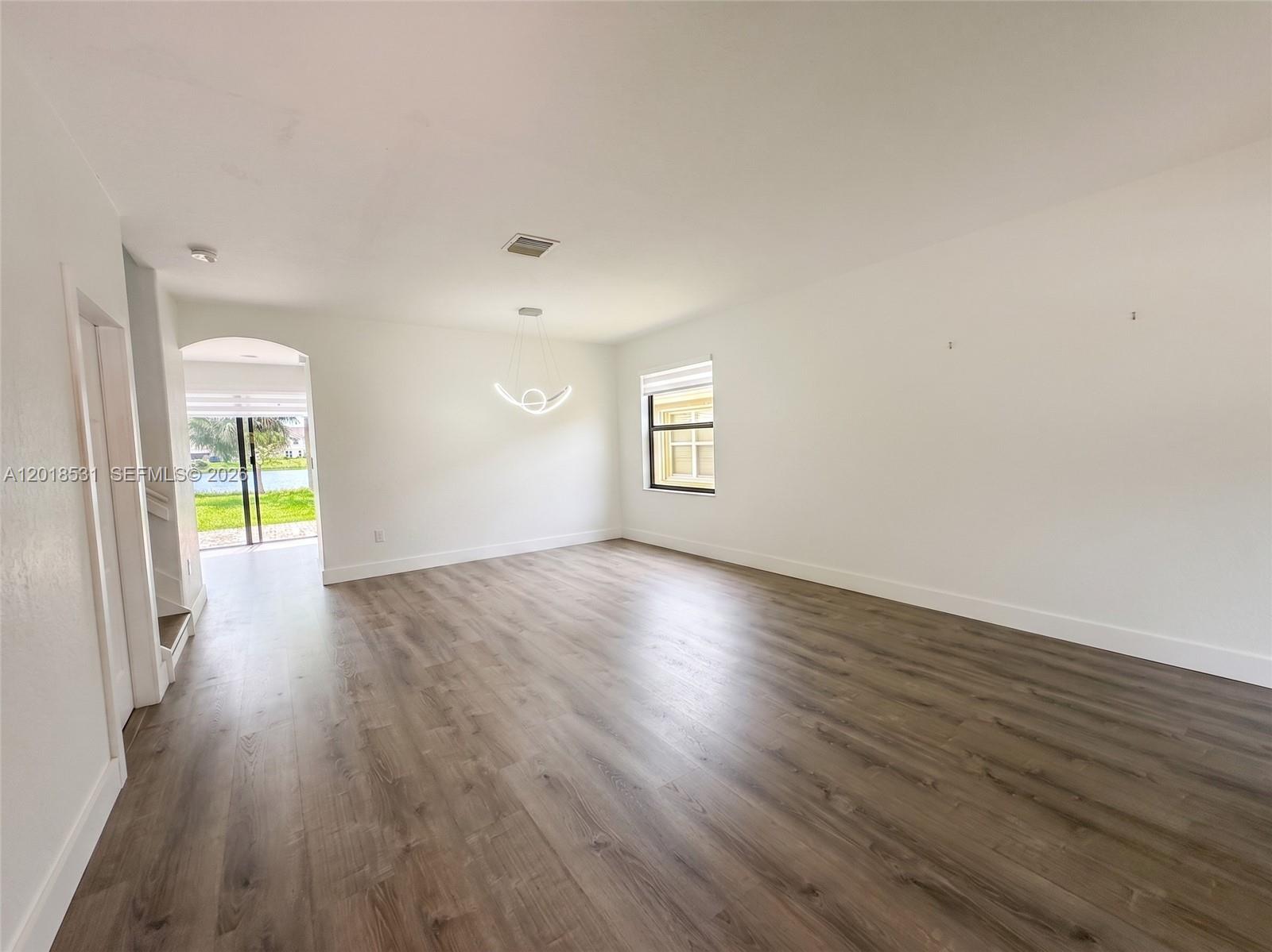 An empty living room features wood-look flooring, white walls, a modern light fixture, and a window — 1575 SE 20th Rd, Homestead FL 33035, HOMESTEAD, FL