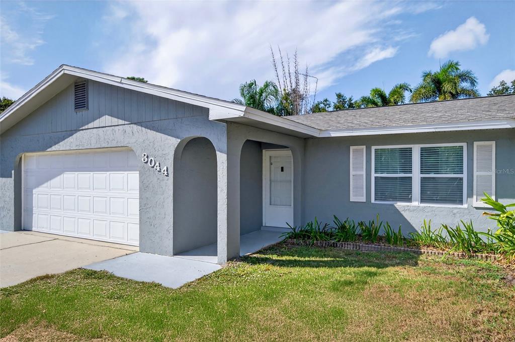 A gray, single-story house features a white garage door, arched entryway, and a front lawn — 3044 GOLDEN RAIN DRIVE, SARASOTA, FL