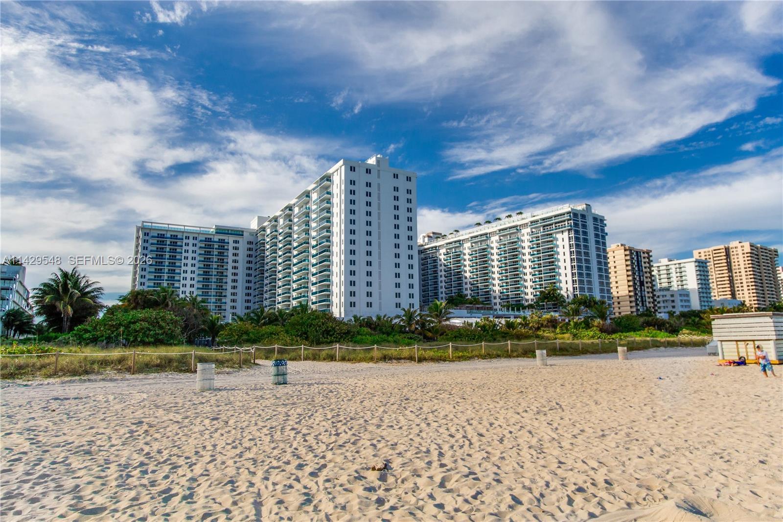 A balcony with a daybed and wooden table overlooks a beach and swimming pool — 102 24th St # 1006, Miami Beach FL 33139, MIAMI BEACH, FL