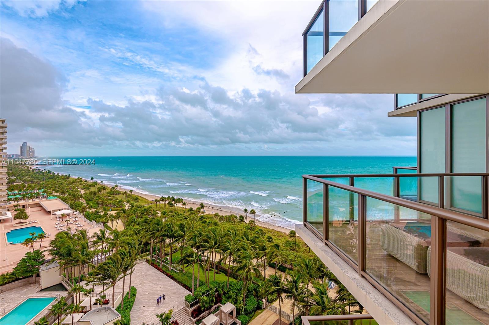 A high-angle view from a balcony overlooks a sandy beach, palm trees, and the ocean — 9703 Collins Ave # 1001, Bal Harbour FL 33154, BAL HARBOUR, FL