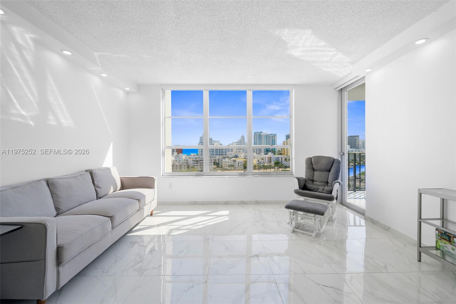 A bright living room features white marble floors, a grey sofa, and a city view — 4101 Pine Tree Dr # 919, Miami Beach FL 33140, MIAMI BEACH, FL