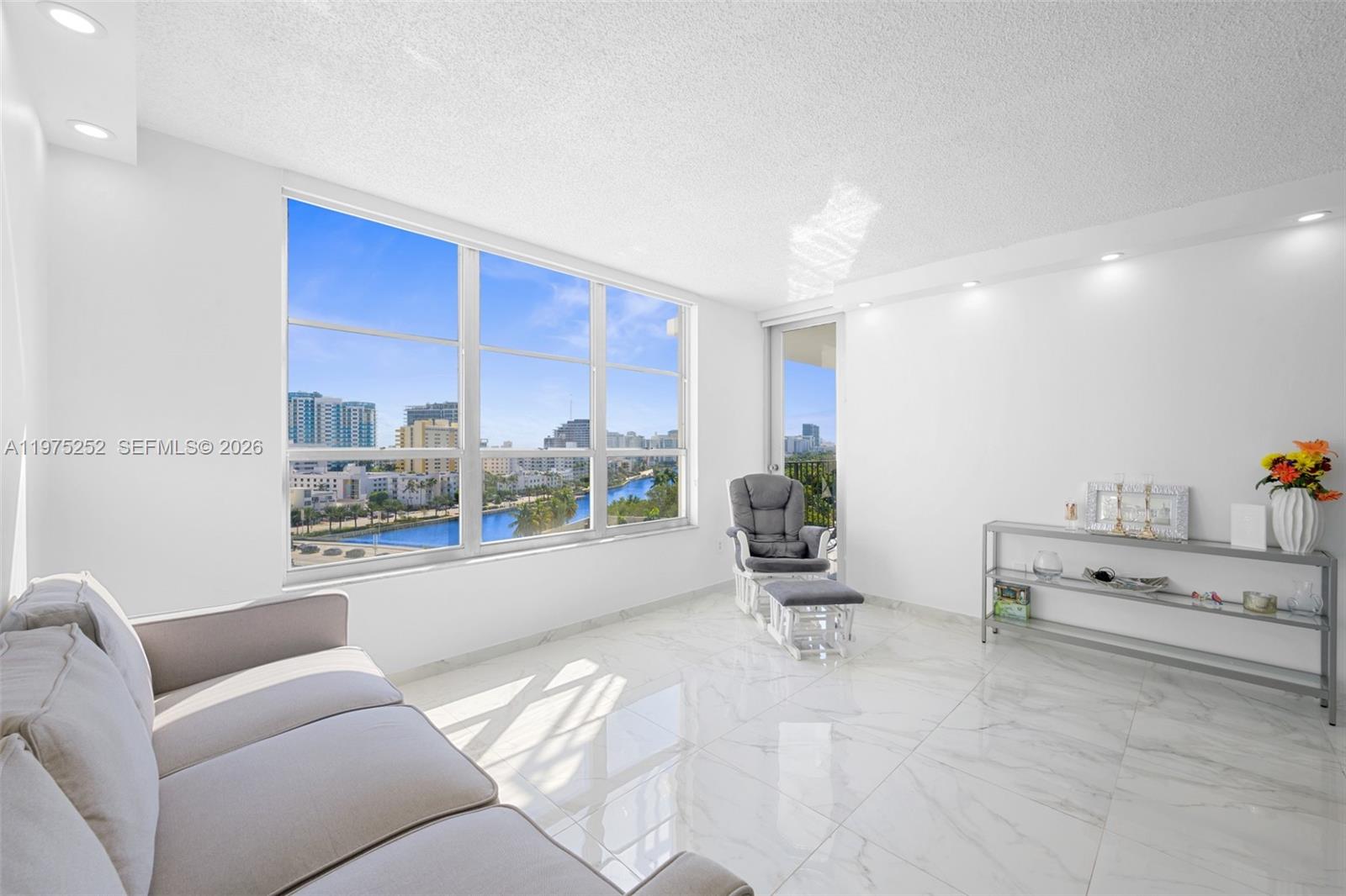 A bright living room features white marble floors, a grey sofa, and large windows overlooking water — 4101 Pine Tree Dr # 919, Miami Beach FL 33140, MIAMI BEACH, FL