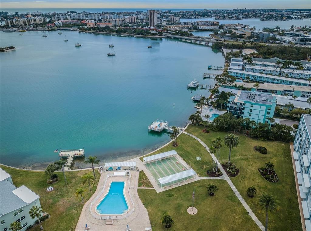 An aerial view shows a swimming pool, tennis courts, and a pier along the water — 1868 SHORE DRIVE S UNIT 102, SOUTH PASADENA, FL