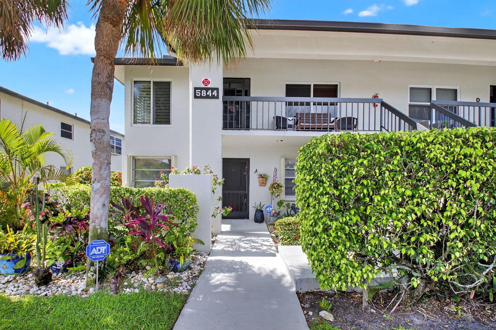 A two-story apartment building exterior features a concrete walkway leading to a ground-floor entrance — 5844 Areca Palm Ct., Unit B, Delray Beach FL 33484, DELRAY BEACH, FL