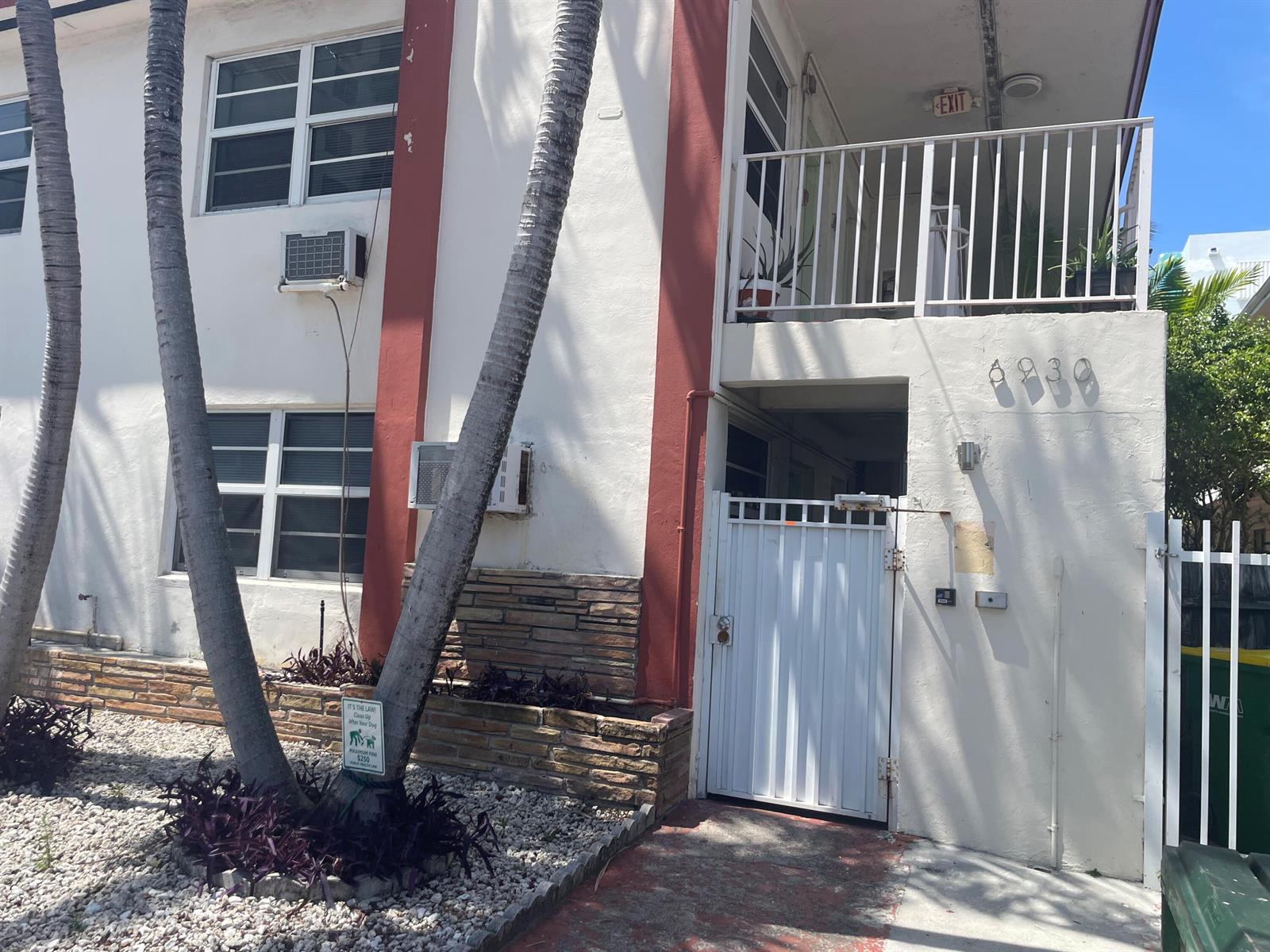 The exterior of a two-story apartment building features white walls, red accents, and palm trees — MIAMI BEACH, FL