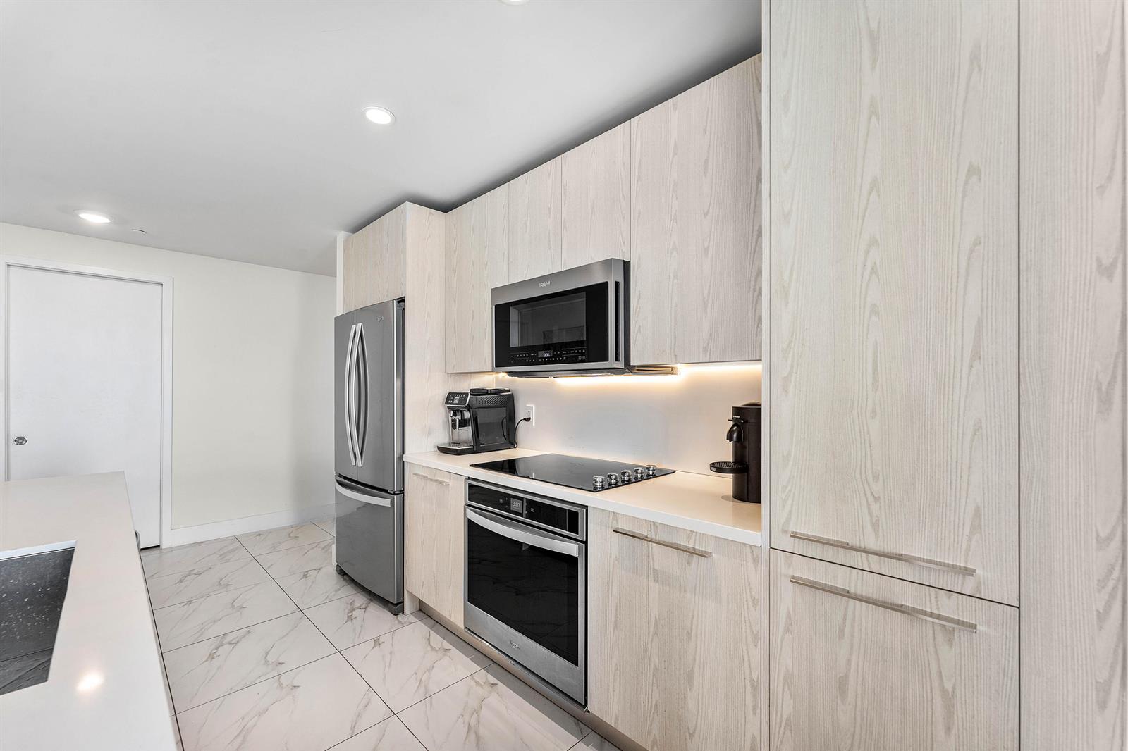 A modern kitchen features light wood cabinetry, stainless steel appliances, and white marble-patterned tile floors — 2000 Metropica Way # 1509, Sunrise FL 33323, SUNRISE, FL