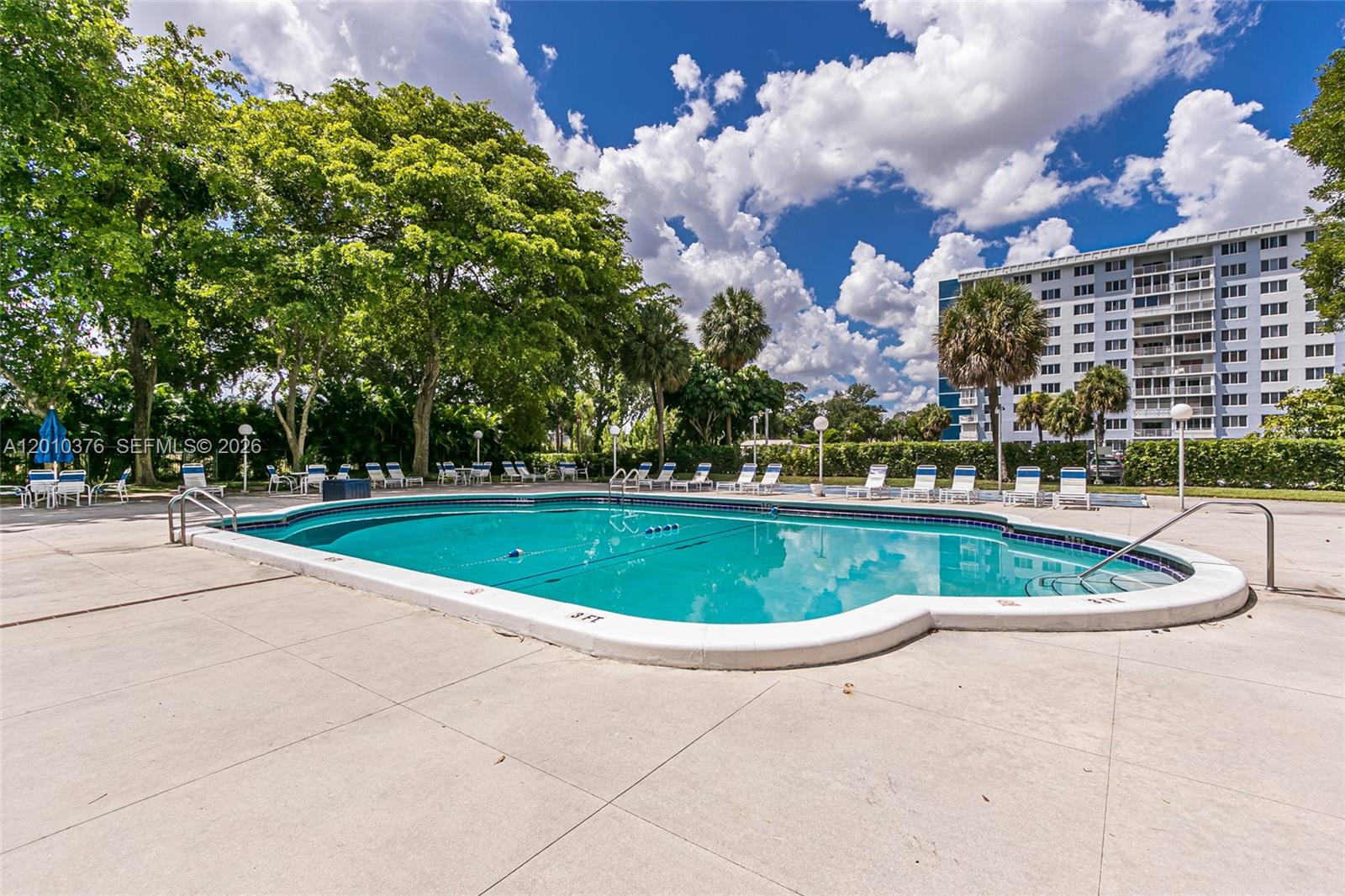 An outdoor swimming pool is surrounded by lounge chairs, trees, and a multi-story apartment building — 3800 Hillcrest Dr # 1211, Hollywood FL 33021, HOLLYWOOD, FL