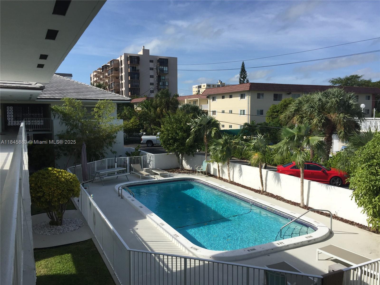 An elevated view of a rectangular swimming pool surrounded by a white fence and palm trees — 201 Sunrise Drive # 102, Key Biscayne FL 33149, KEY BISCAYNE, FL