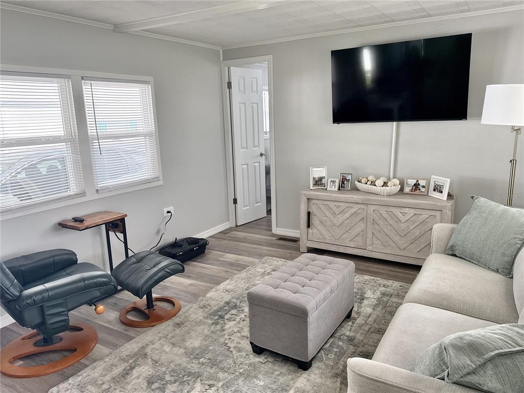 A living room features a wall-mounted television, a grey sofa, an armchair, and wood-look flooring — 7214 MOUNT FAIRFIELD ROAD NE UNIT 527, ST PETERSBURG, FL