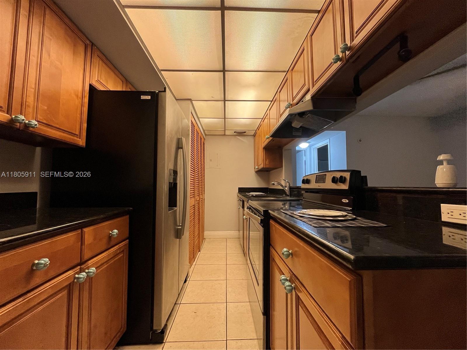 A galley kitchen features wood cabinets, black countertops, stainless steel appliances, and a drop ceiling — 4469 Treehouse Ln # B, Tamarac FL 33319, TAMARAC, FL
