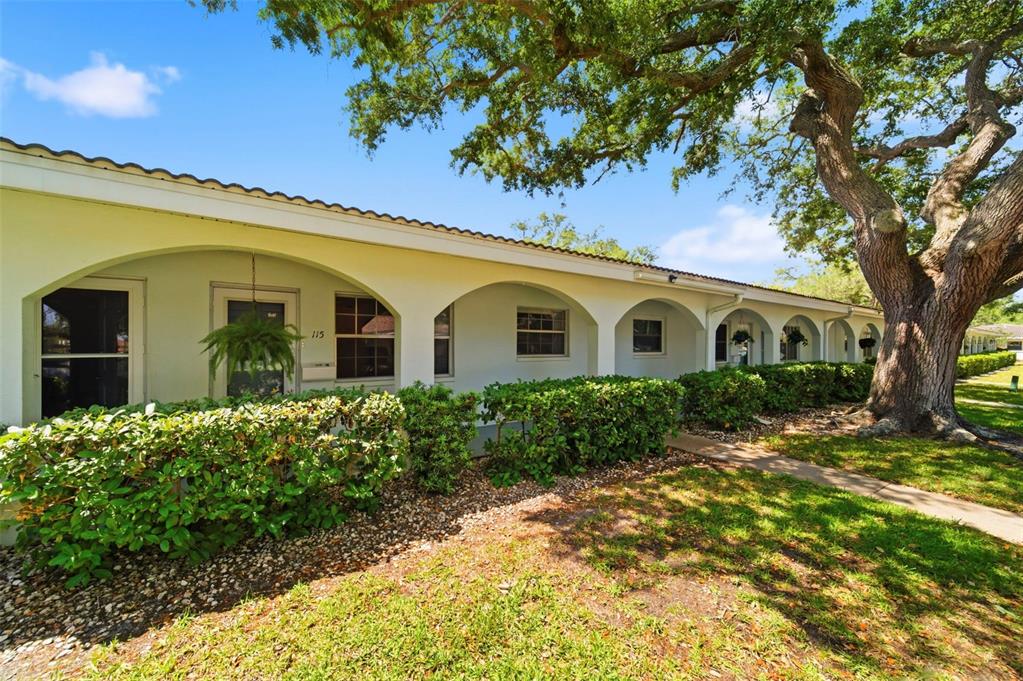 A light-colored, single-story building features a covered walkway with arched openings and mature trees — 11200 102ND AVENUE UNIT 115, SEMINOLE, FL