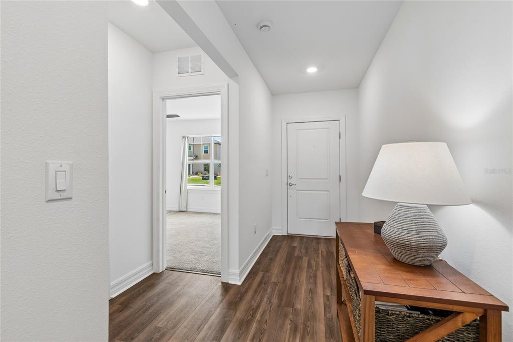 A hallway with wood-look flooring leads to an entryway door and an adjacent carpeted room — 3883 CAPRI COAST DRIVE, PLANT CITY, FL
