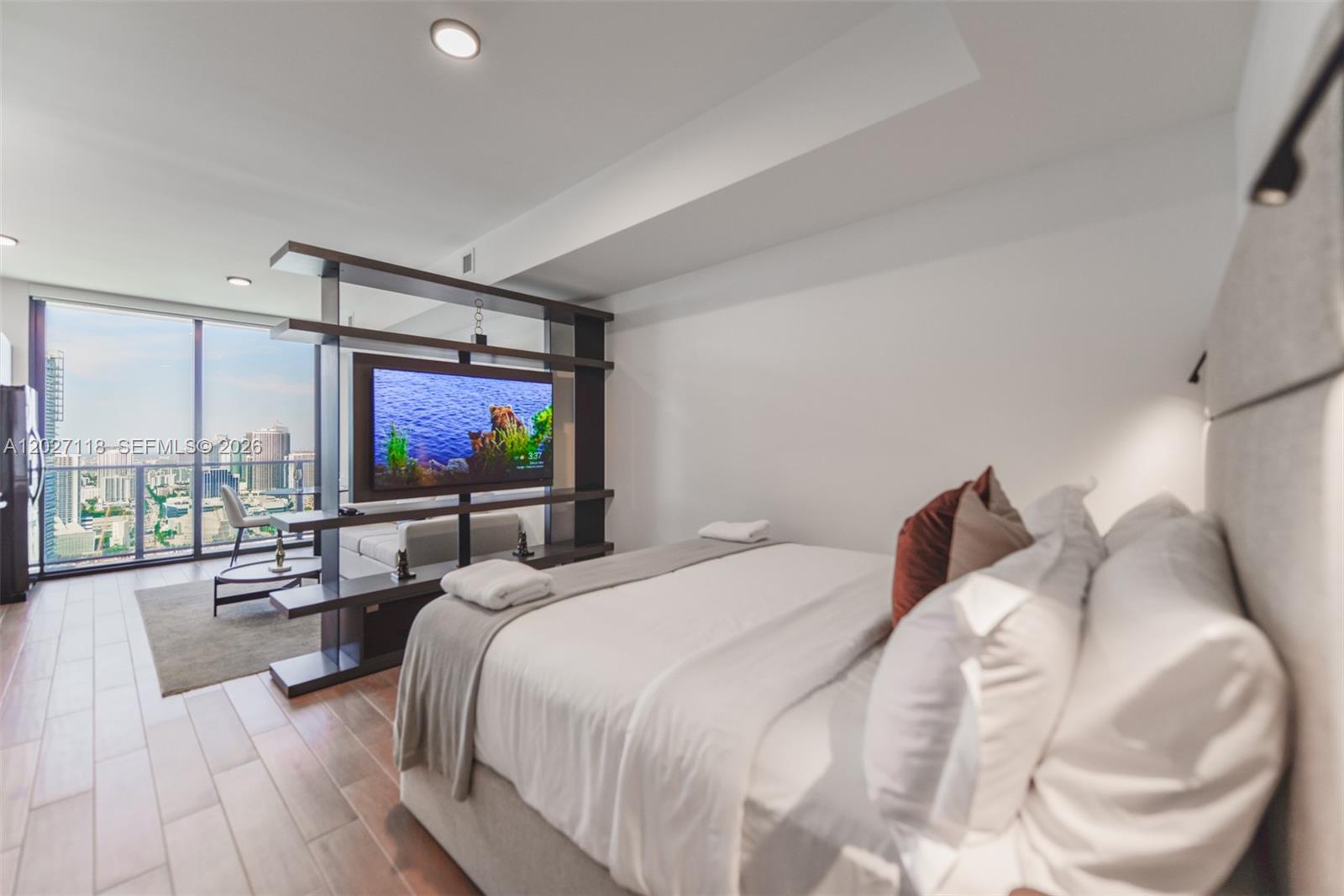 A bedroom features a bed, a room-dividing shelving unit with a television, and floor-to-ceiling windows — 398 NE 5th St # 3505, Miami FL 33132, MIAMI, FL