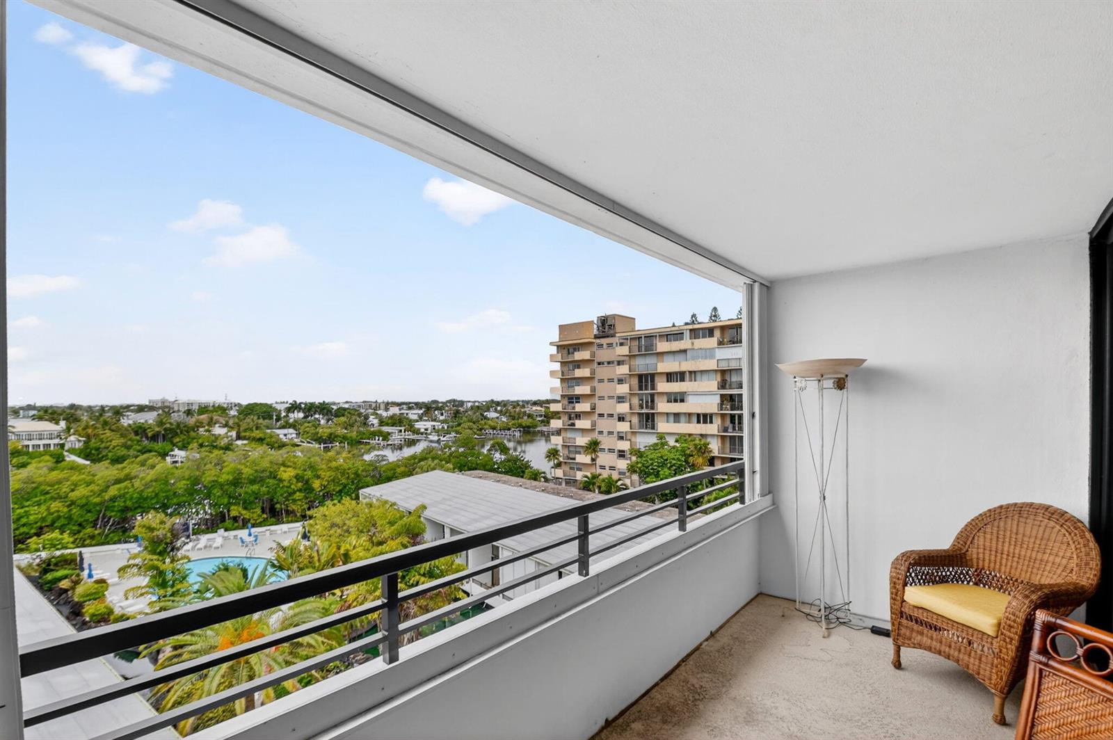 A balcony with a wicker chair overlooks a swimming pool, lush trees, and nearby buildings — 3589 S Ocean Boulevard # 609, South Palm Beach FL 33480, SOUTH PALM BEACH, FL