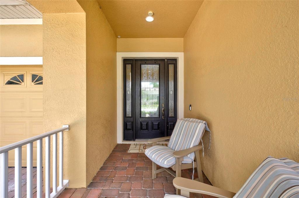 A covered front porch with brick flooring, two striped chairs, and a dark glass-paneled door — 1801 BURDOCK DRIVE, WINTER GARDEN, FL