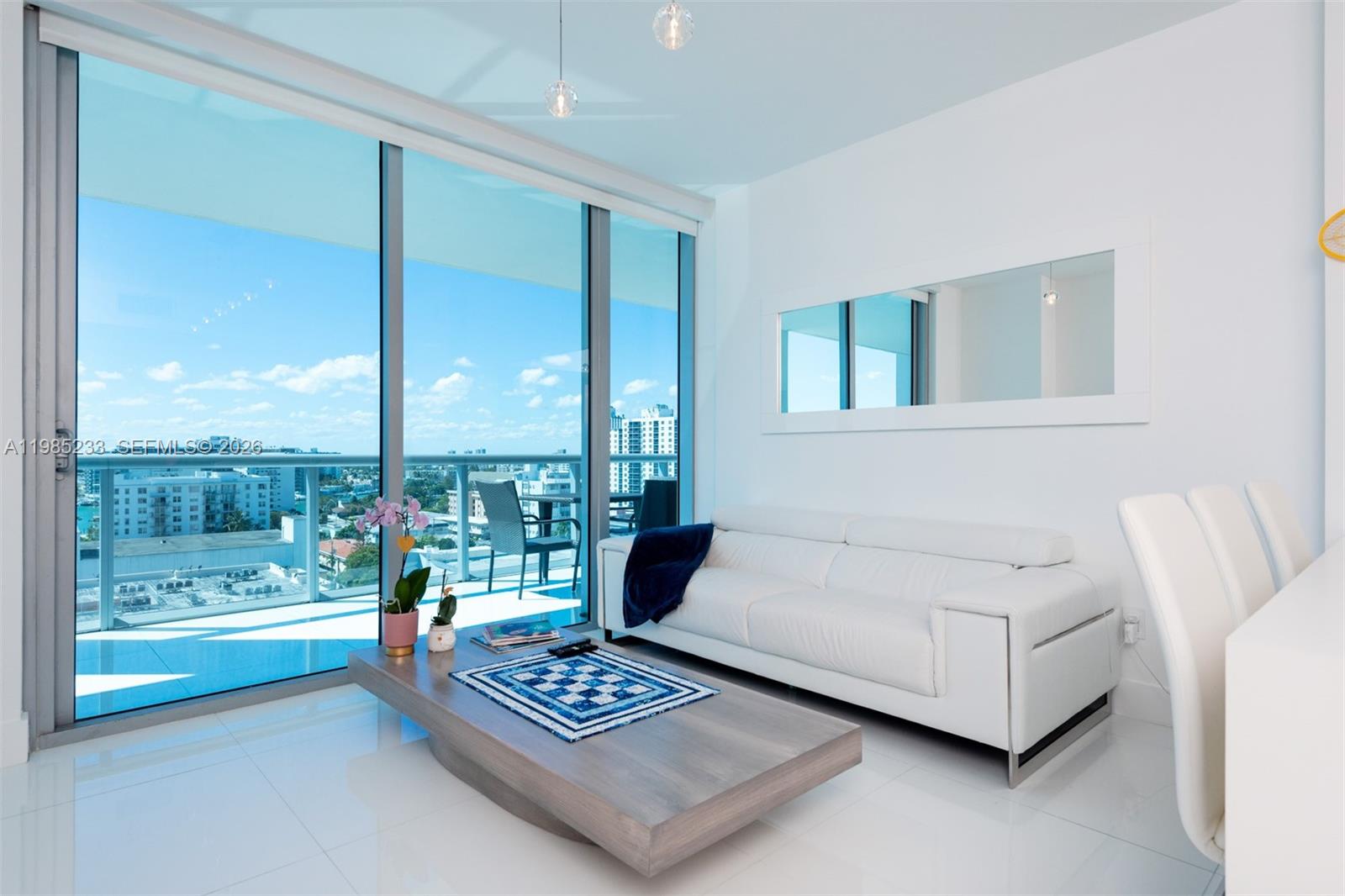 A modern living room features a white sofa, a low wooden coffee table, and floor-to-ceiling windows — 6799 Collins Ave # 906, Miami Beach FL 33141, MIAMI BEACH, FL