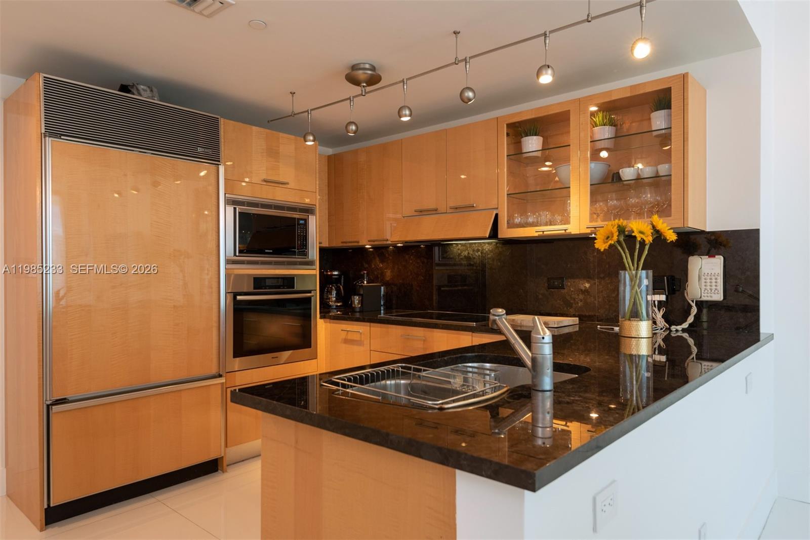 A kitchen features light wood cabinetry, black granite countertops, stainless steel appliances, and track lighting — 6799 Collins Ave # 906, Miami Beach FL 33141, MIAMI BEACH, FL