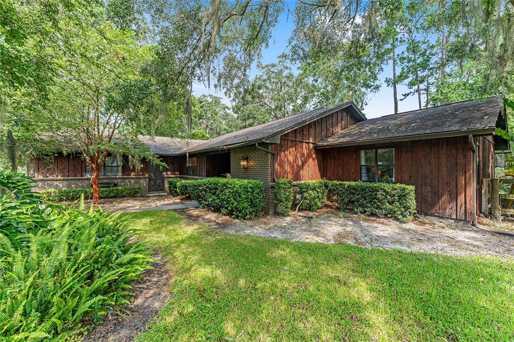 A single-story house with dark wood siding and a brick base sits among mature trees — 8101 NW 46TH STREET, OCALA, FL
