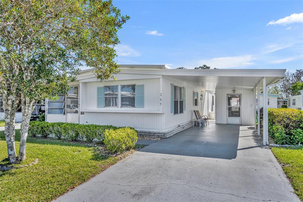 A white manufactured home features a carport, blue shutters, and a landscaped front yard — 269 KELOU COURT, LEESBURG, FL