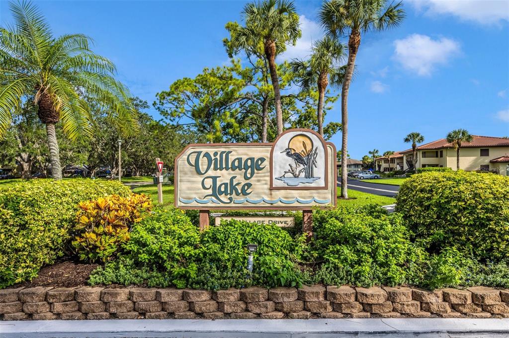 A sign for Village Lake stands in front of lush landscaping and palm trees — 5178 LAKE VILLAGE DRIVE UNIT 86, SARASOTA, FL