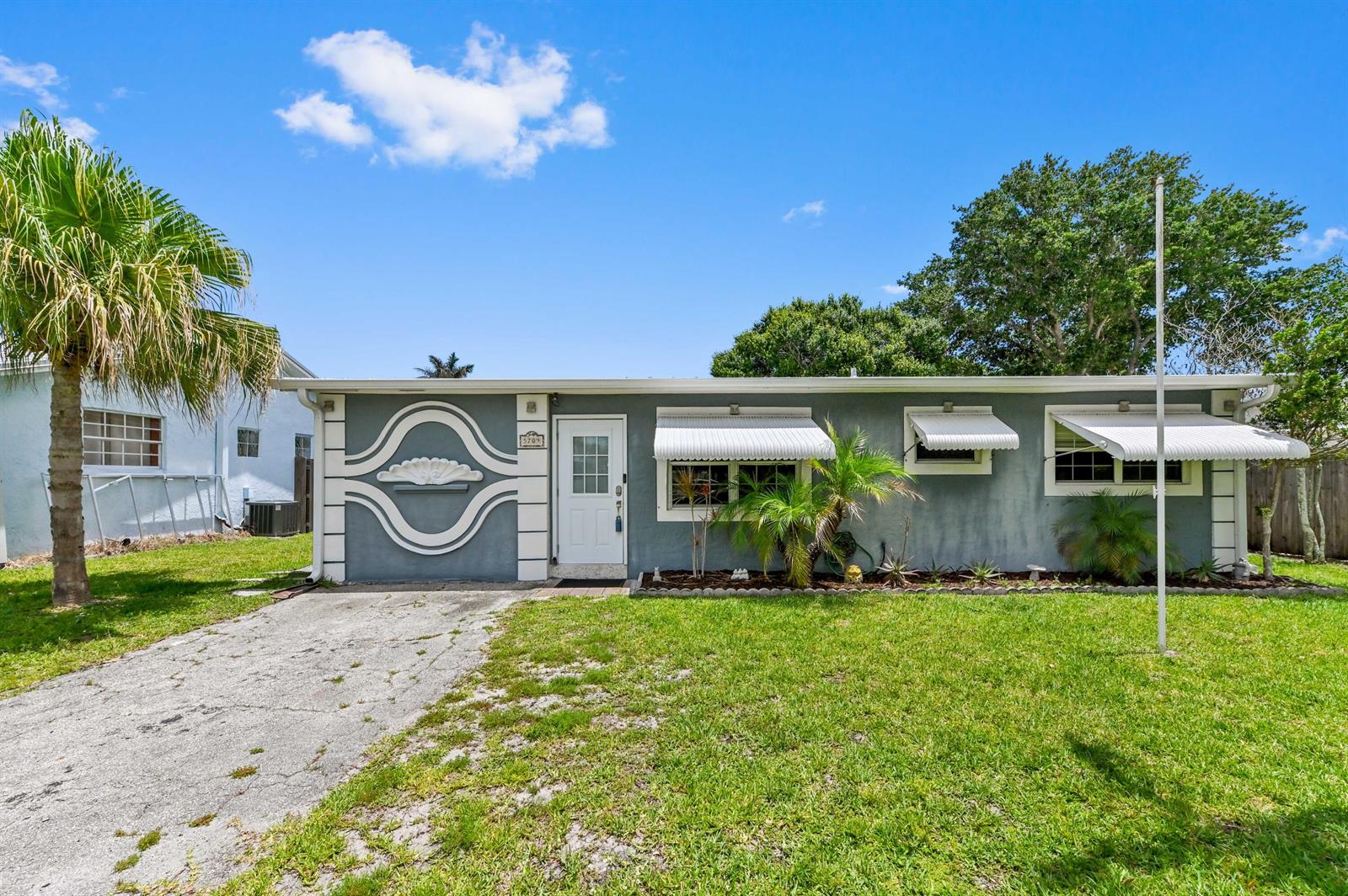 A gray single-story house features a white decorative facade, metal awnings, and a concrete driveway — 5709 SE Indigo Avenue, Stuart FL 34997, STUART, FL