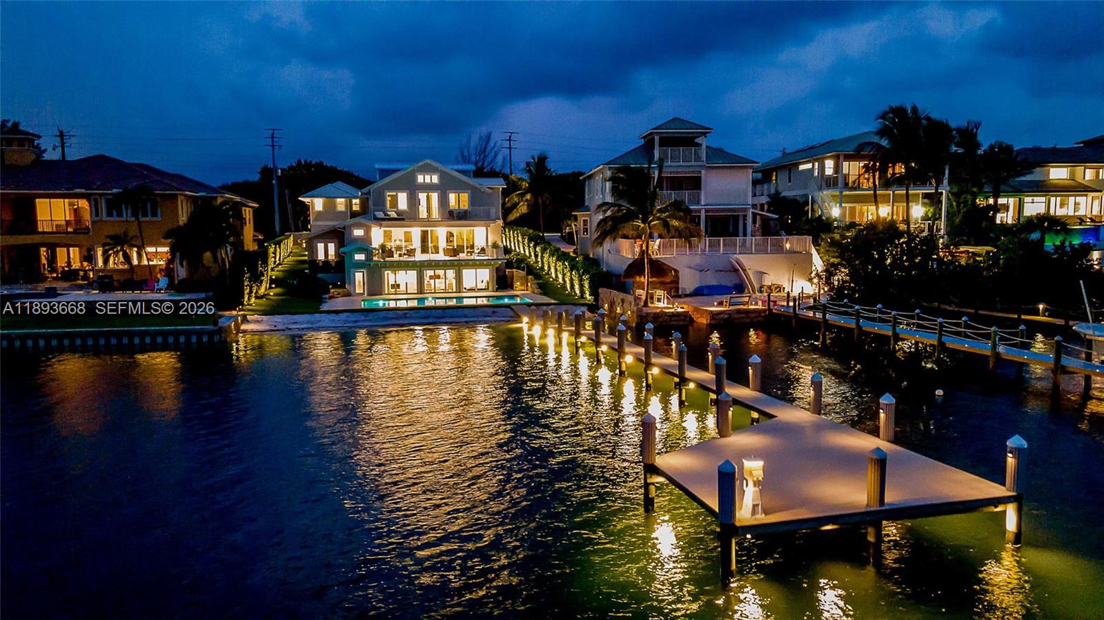 An illuminated waterfront house features a long wooden dock extending into the canal at dusk — 18119 SE Federal Hwy, Jupiter FL 33469, JUPITER, FL
