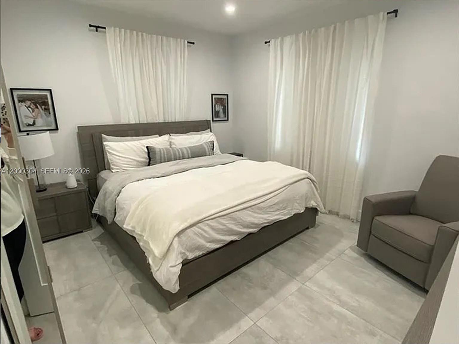 A bedroom features a gray bed, matching nightstand, armchair, and white curtains on tiled floors — 1935 SW 24th Ter, Miami FL 33145, MIAMI, FL
