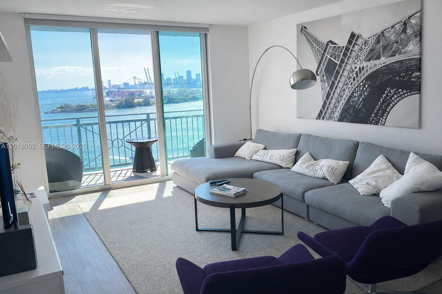 A living room features a gray sectional sofa, purple armchairs, and a balcony with water views — 90 Alton Rd # 1804, Miami Beach FL 33139, MIAMI BEACH, FL