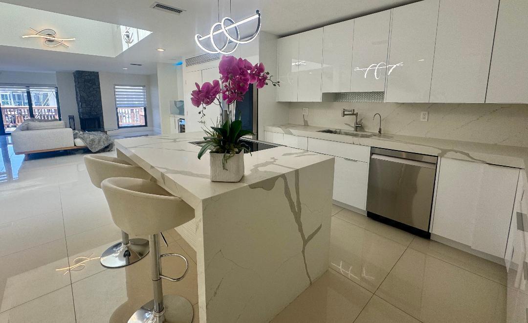 A modern kitchen features a marble waterfall island, white cabinetry, and stainless steel appliances — 1127 Boca Cove Lane, Highland Beach FL 33487, HIGHLAND BEACH, FL