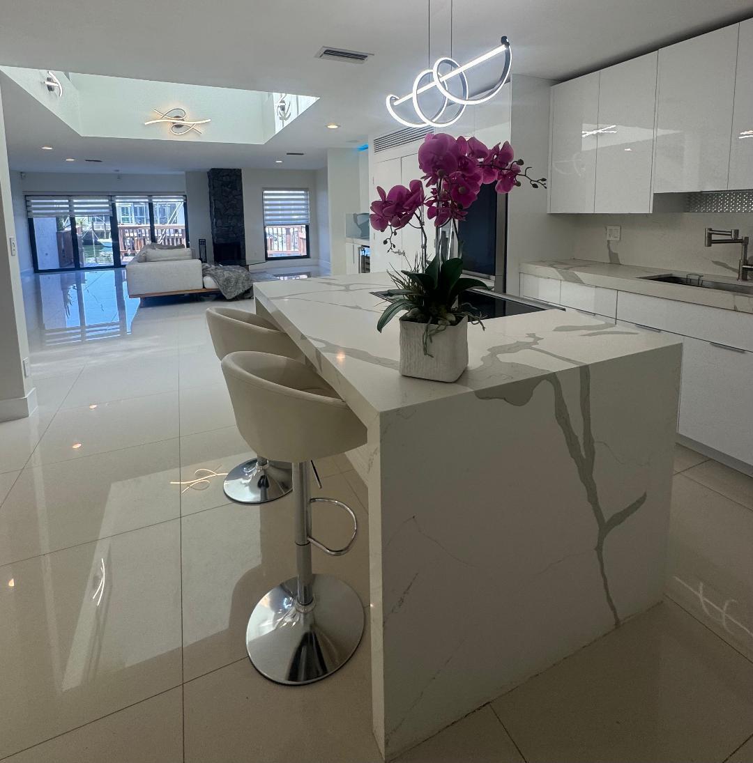 A modern kitchen island with a waterfall quartz countertop sits before an open-concept living area — 1127 Boca Cove Lane, Highland Beach FL 33487, HIGHLAND BEACH, FL
