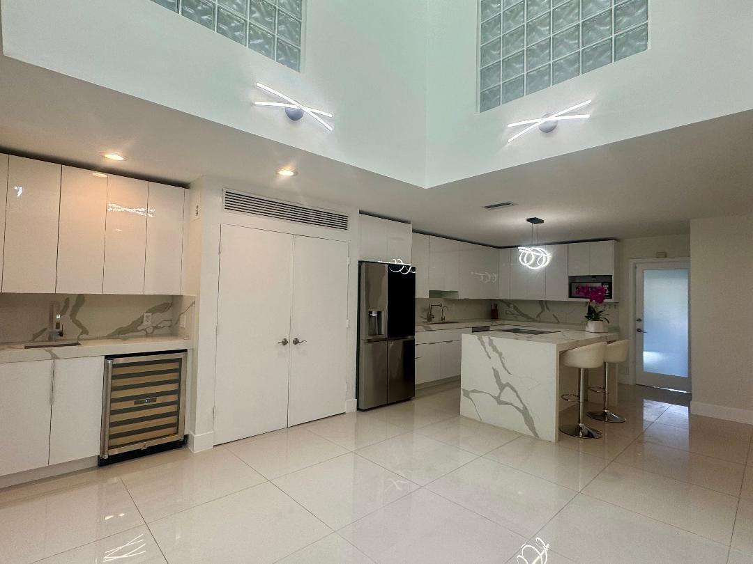 A modern kitchen features white glossy cabinets, marble-patterned countertops, a center island, and glass blocks — 1127 Boca Cove Lane, Highland Beach FL 33487, HIGHLAND BEACH, FL