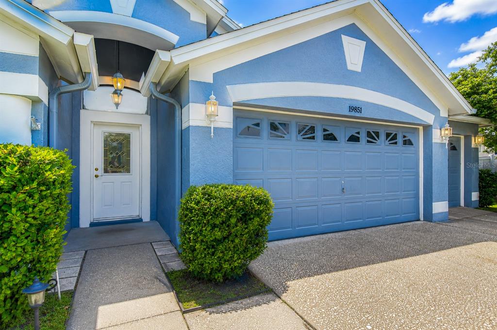 A blue house exterior features a white front door, a garage, and manicured green bushes — 19851 ELLENDALE DRIVE, LAND O LAKES, FL