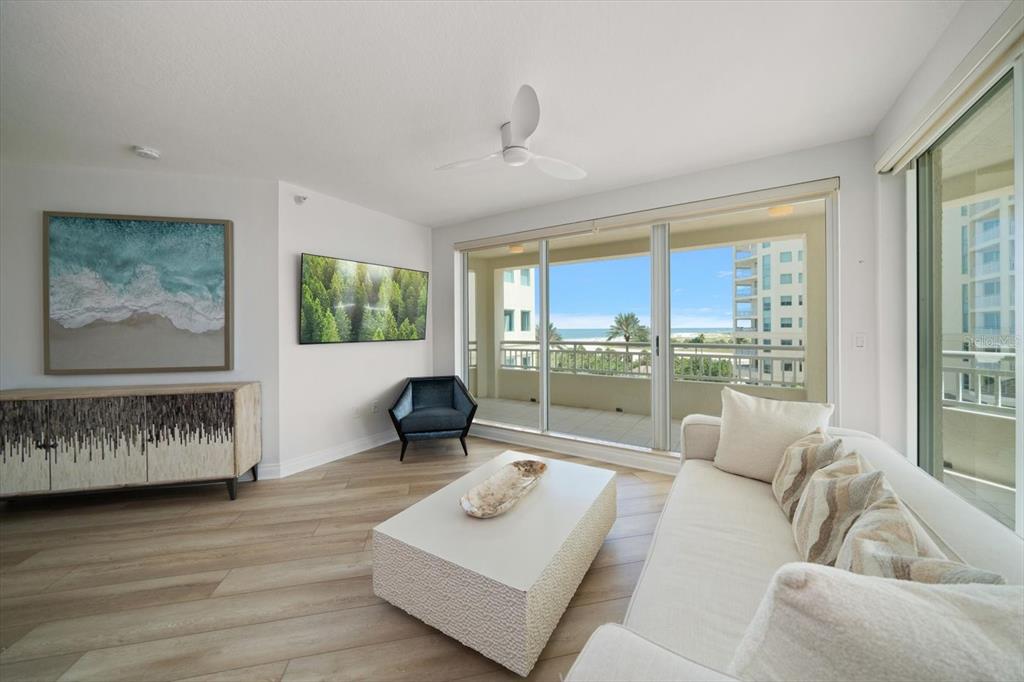 A bright living room features light wood floors, a white sofa, and a balcony view — 1180 GULF BLVD UNIT 504, CLEARWATER BEACH, FL